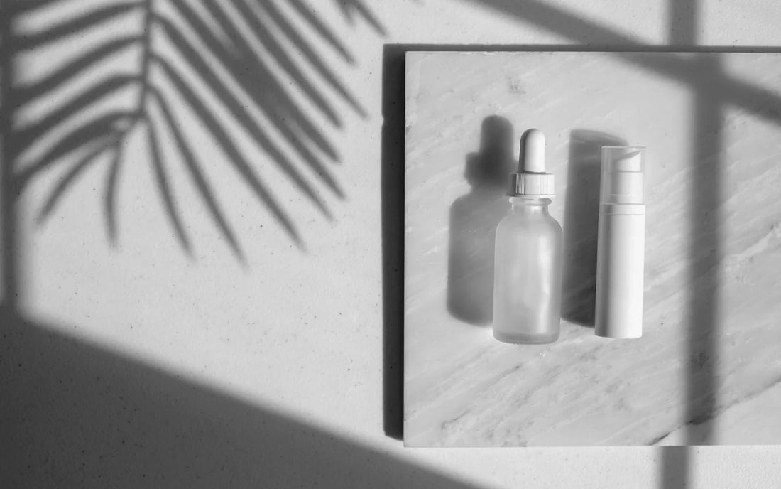 A glass dropper bottle and a white pump bottle on a marble surface, with a palm leaf shadow.