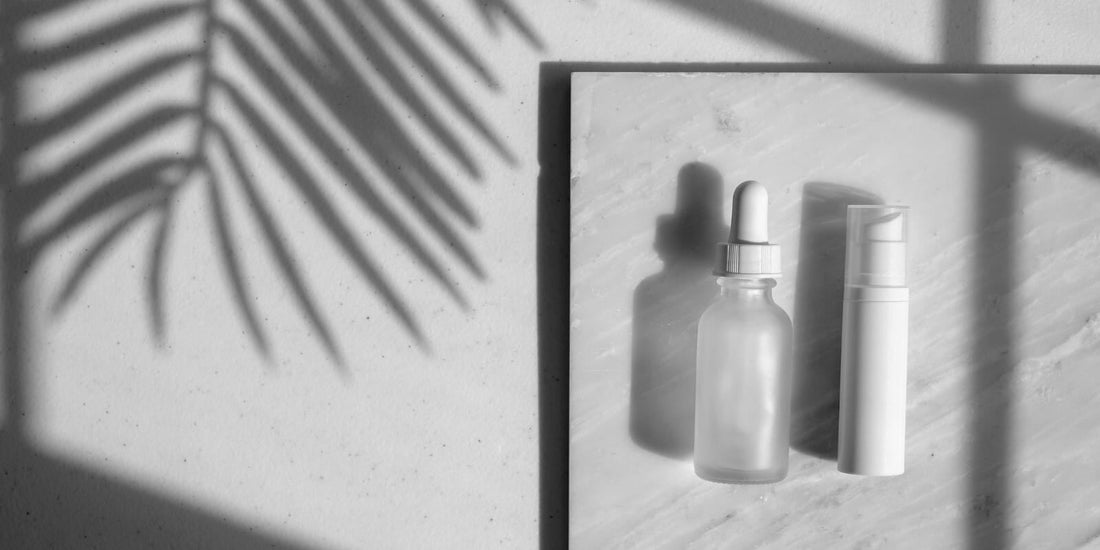 Minimalist skincare flat lay with a frosted glass dropper bottle and a white pump bottle on a marble slab, accented by shadows of palm leaves.