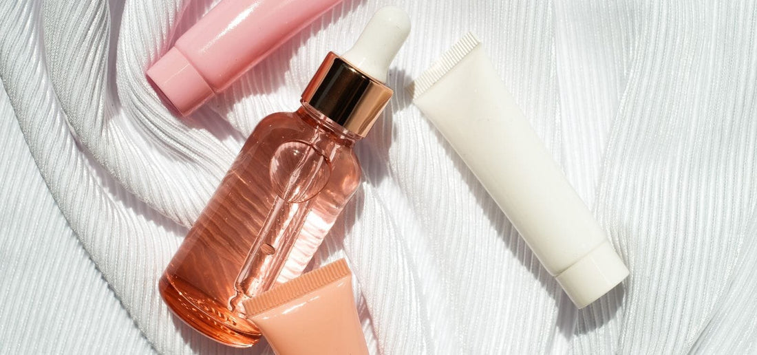 Flat lay of Indigo Private Label skincare on white fabric: rose-gold dropper, white tube, and two soft pink tubes.