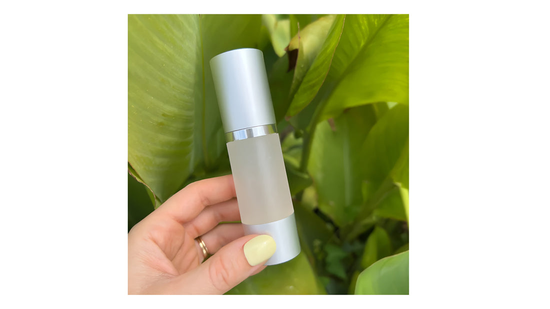Hand holding an Indigo vitamin C serum bottle with a silver cap, frosted body, and lush green leaves in the background.