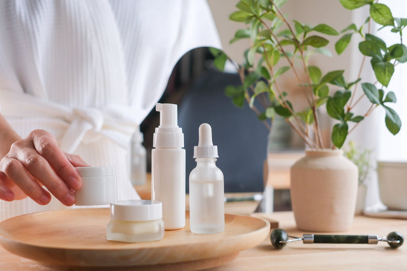 A person in a white robe holding a skincare jar next to minimalist cosmetic bottles on a wooden tray, with a jade roller and potted plant in the background.