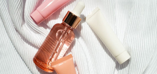 Flat lay of Indigo Private Label skincare on white fabric: rose-gold dropper, white tube, and two soft pink tubes.