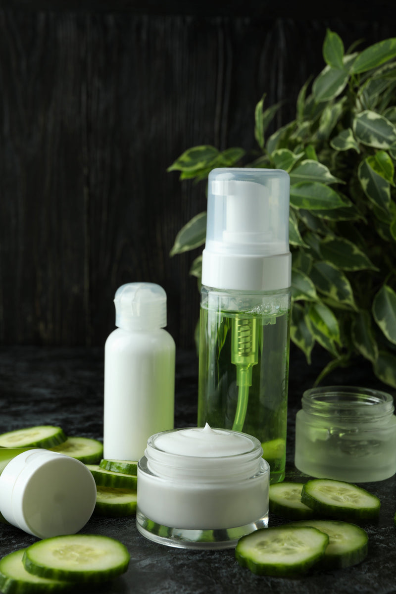 Close-up of skincare products with cucumber slices, including a white bottle, a foaming cleanser, and cream jars.