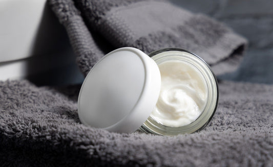 Open glass jar of white face cream on a soft gray towel, with lid resting to the side in a bathroom setting.